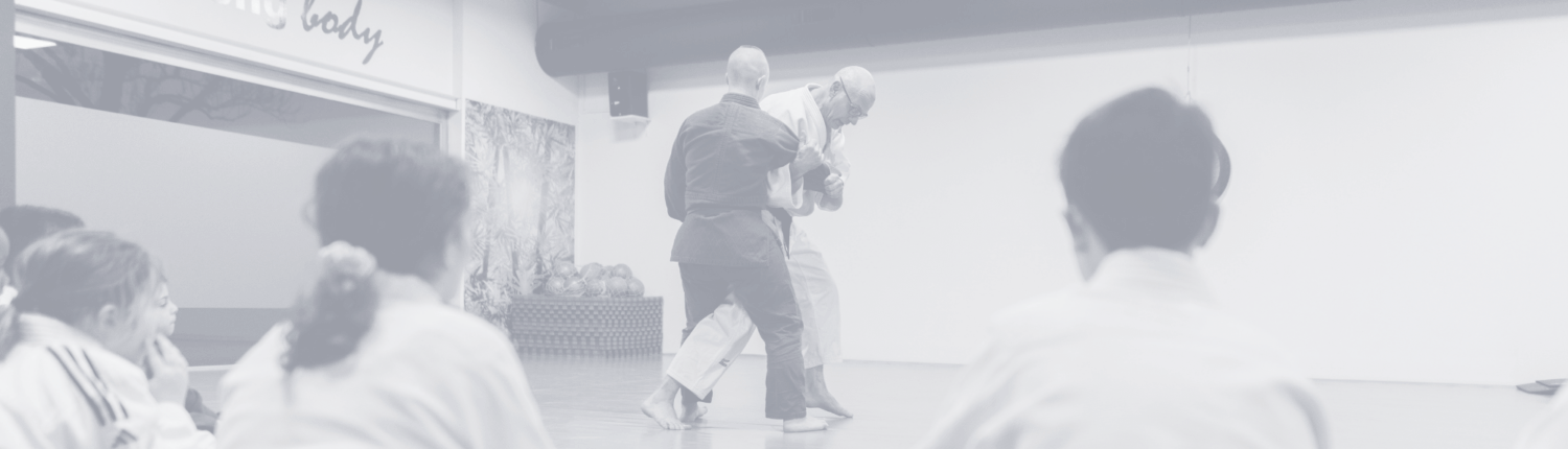 een groep kinderen die zitten en kijken hoe 2 mensen judoen